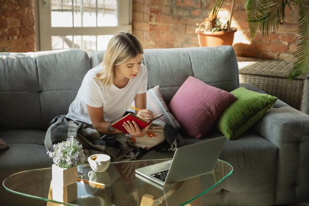 Obteniendo tareas. Mujer caucásica, autónoma durante el trabajo en la oficina en casa mientras está en cuarentena. Joven empresaria en casa, auto aislado. Usando gadgets. Trabajo remoto, prevención de la propagación del coronavirus.