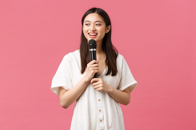 Ocio, emociones de las personas y concepto de estilo de vida. Alegre niña asiática sonriente en karaoke, disfrutando de los fines de semana, cantando canciones en el micrófono, realizando stand-up, fondo rosa de pie.