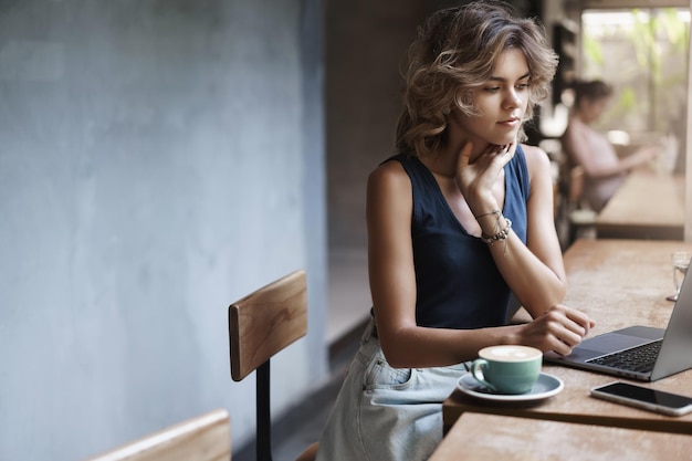 Ocupada, atractiva, joven y moderna mujer de negocios, preparar proyecto de lectura, artículo, sentarse cerca de la ventana, mesa de café, beber café, pensar, mirar, computadora portátil, pantalla, estudiante, tutor, hacer, arreglos, futuro, lección