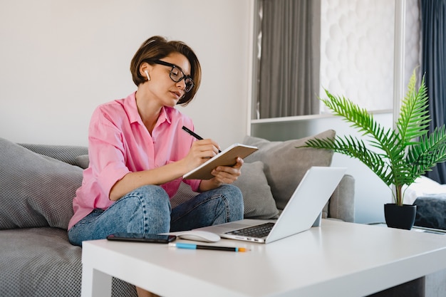 Ocupada mujer seria en camisa rosa sentada concentrada haciendo notas pagando facturas en el sofá en casa en la mesa trabajando en línea en la computadora portátil desde casa