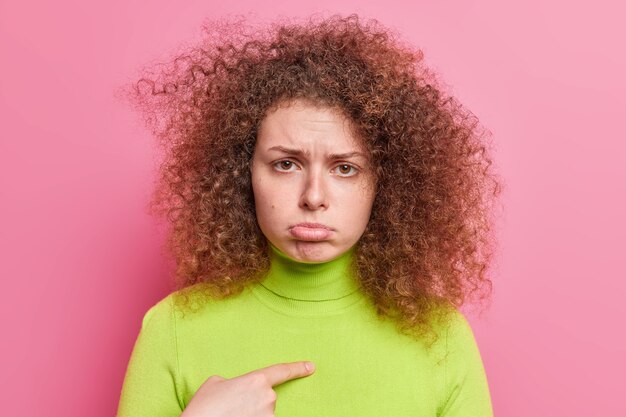 Ofendida, insultada, bonita adolescente con cabello rizado y tupido apunta a sí misma monederos labio inferior tiene expresión disgustada usa cuello alto verde aislado sobre una pared rosa pregunta ¿me culpas