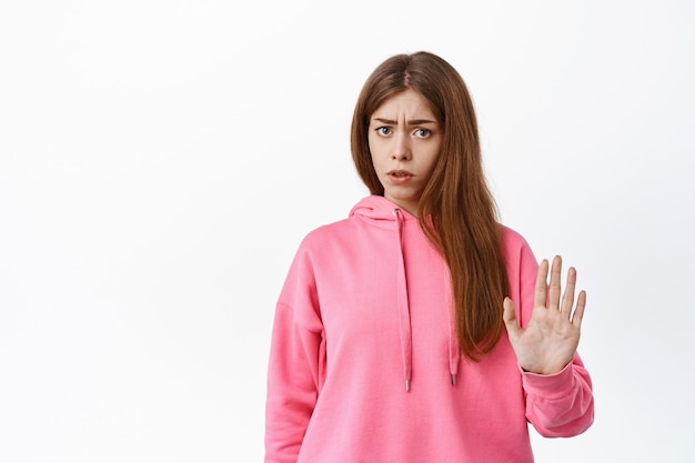 Oferta de bloqueo de mujer joven disgustada, rechazando algo malo, mostrando gesto de bloqueo de parada, rechazando, de pie sobre una pared blanca