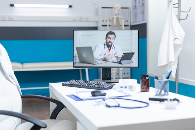 Oficina médica vacía con un médico remoto que tiene una discusión en línea durante la conferencia de reunión de videollamada de telemedicina. Computadora con llamada de videoconferencia de teletrabajo en pantalla. Apoyo a los servicios de medicina