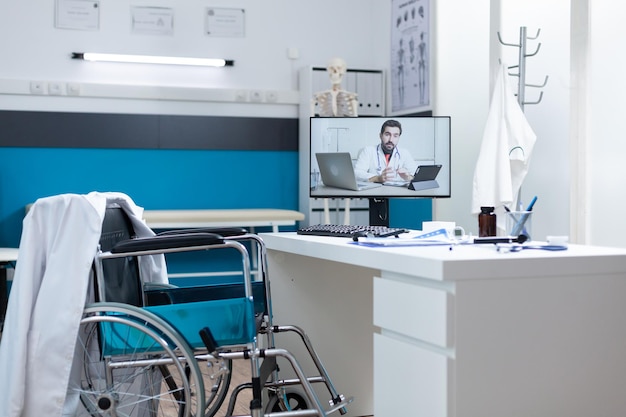 Oficina de médicos vacía con médico remore hablando durante la conferencia de videollamada en línea en la pantalla de la computadora. Habitación de hospital equipada con herramientas médicas profesionales. Servicios de medicina