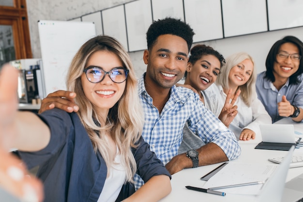 Oficinista negro en camisa a cuadros abrazando secretaria rubia mientras ella hace selfie. Jóvenes directivos de empresa internacional divirtiéndose durante la reunión.