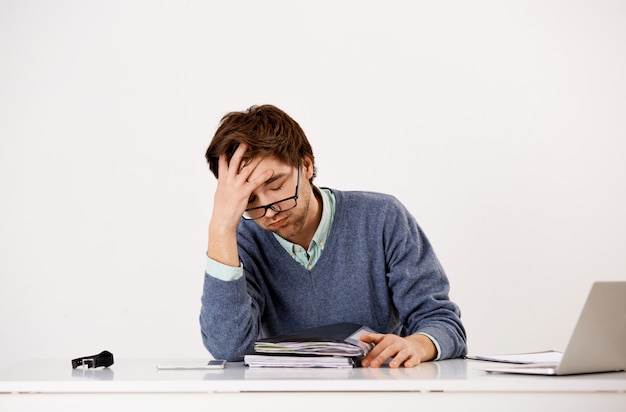 Oficinista de sexo masculino cansado, suspirando incómodo, trabajando hasta tarde, tiene plazos, estudiando informes y documentos como sentarse a la mesa con la computadora portátil, con la cara palmada angustiada