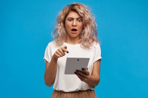 Oh Dios mío. Chica estudiante asombrada de ojos de insecto con ropa elegante sosteniendo una computadora táctil y apuntando a la pantalla con la boca abierta