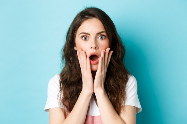 Oh Dios mío. Ciérrese encima del retrato de la cara de la niña sorprendida, tocando la cabeza y mirando a la cámara sorprendida, escuche malas noticias, de pie sobre fondo azul.