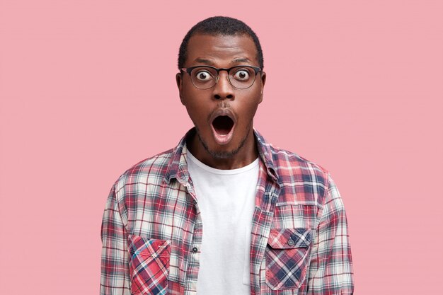 Oh Dios mío. Hombre afroamericano de piel oscura sorprendido mira fijamente a la cámara con expresión de asombro, usa gafas y camisa a cuadros