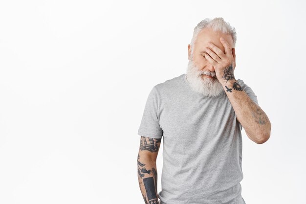 Oh Dios mío. Hombre mayor cansado y molesto con la palma de la mano en el rostro angustiado o agotado, de pie irritado en una camiseta gris informal contra el fondo blanco