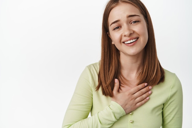 Oh, gracias, eso es tan dulce. Cara de mujer joven tocada y halagada, tome la mano en el pecho y sonría con gratitud, exprese aprecio, agradezca, de pie contra el fondo blanco