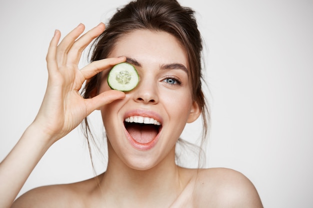 Ojo de ocultación sonriente de la muchacha desnuda hermosa joven detrás de la rebanada del pepino que mira la cámara sobre el fondo blanco.