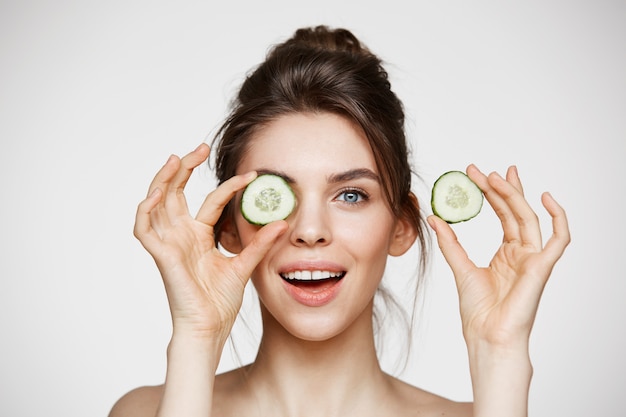 Ojo de ocultación sonriente de la muchacha desnuda hermosa joven detrás de la rebanada del pepino que mira la cámara sobre el fondo blanco.