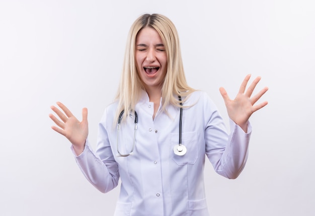 Con los ojos cerrados, alegre, doctor, joven, rubio, niña, llevando, estetoscopio, y, médico, bata, en, aparato dental, levantado, manos, aislado