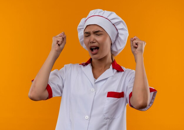 Con los ojos cerrados, alegre joven cocinera vistiendo uniforme de chef mostrando sí gesto sobre fondo naranja aislado