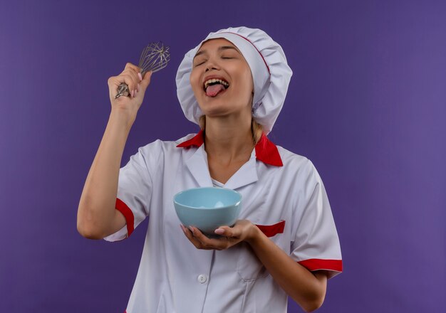 Con los ojos cerrados alegre joven cocinera vistiendo uniforme de chef sosteniendo un batidor y un cuenco mostrando la lengua sobre fondo aislado