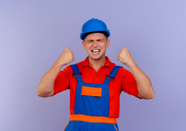 Con los ojos cerrados alegre joven constructor vistiendo uniforme y casco de seguridad mostrando sí gesto en púrpura