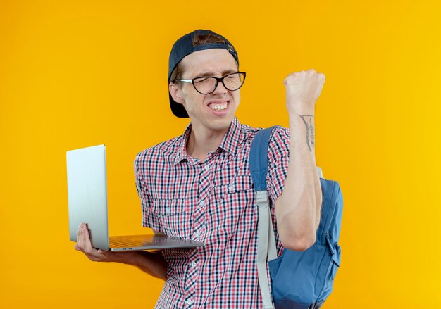 Con los ojos cerrados, alegre, joven estudiante muchacho con bolso trasero y gafas y gorra sosteniendo un portátil mostrando gesto sí en blanco