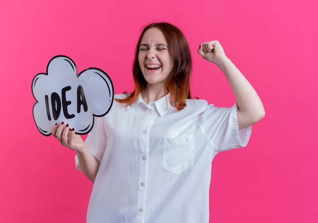 Con los ojos cerrados, alegre joven pelirroja sosteniendo la burbuja de la idea y levantando el puño aislado en rosa