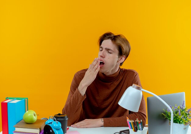 Con los ojos cerrados bostezo joven estudiante sentado en un escritorio con herramientas escolares y la boca cubierta con la mano en amarillo