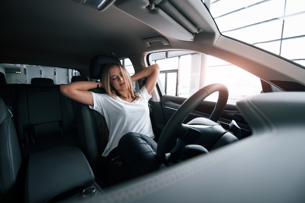 Con los ojos cerrados. Chica en coche moderno en el salón. Durante el día en interiores. Comprar vehículo nuevo