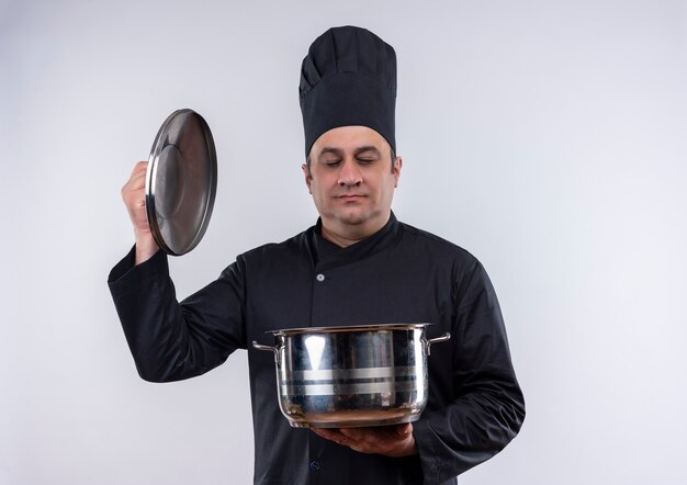 Con los ojos cerrados, cocinero masculino de mediana edad en uniforme de chef sosteniendo una cacerola y tapa