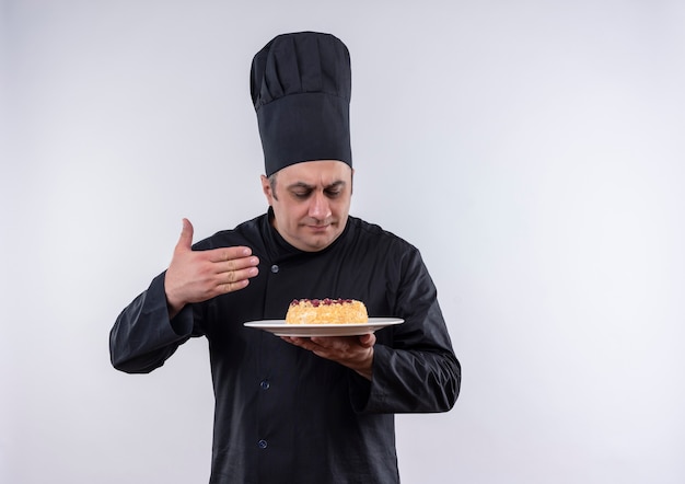 Con los ojos cerrados, cocinero de sexo masculino de mediana edad en uniforme de chef oliendo el pastel en el plato en su mano