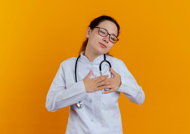 Con los ojos cerrados complacida joven doctora vistiendo bata médica y estetoscopio con gafas poniendo las manos en el corazón aislado