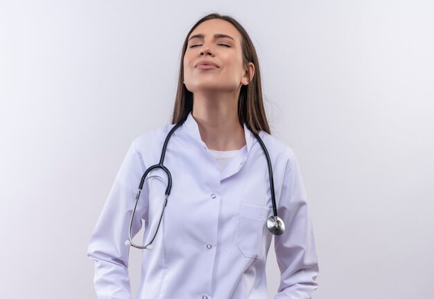 Con los ojos cerrados complacido joven vistiendo estetoscopio bata médica en blanco aislado