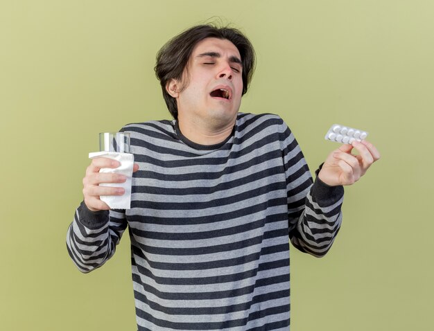 Con los ojos cerrados confundido joven enfermo sosteniendo pastillas y un vaso de agua aislado sobre fondo verde oliva