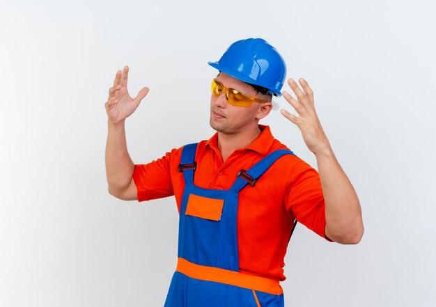 Con los ojos cerrados, el constructor masculino joven cansado con uniforme y casco de seguridad y gafas de seguridad extiende las manos en blanco