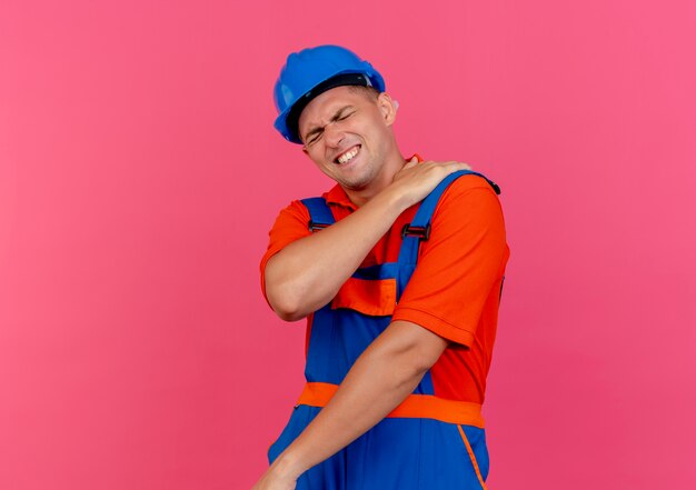 Con los ojos cerrados, el constructor masculino joven enfermo vistiendo uniforme y casco de seguridad poniendo la mano sobre el hombro dolorido en rosa