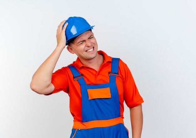 Con los ojos cerrados, el constructor masculino joven preocupado vistiendo uniforme y casco de seguridad poniendo la mano en la cabeza en blanco