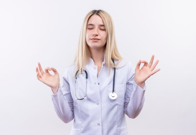 Con los ojos cerrados doctor joven rubia con estetoscopio y bata médica mostrando gesto okey con ambas manos en la pared blanca aislada