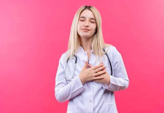 Con los ojos cerrados, el doctor joven rubia con estetoscopio en bata médica puso sus manos en el corazón en la pared rosa aislada