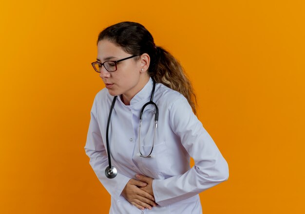 Con los ojos cerrados, doctora joven enferma con bata médica y estetoscopio con gafas agarró dolor de estómago aislado en la pared naranja