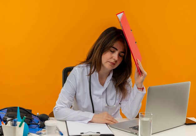 Con los ojos cerrados, doctora de mediana edad cansada con bata médica con estetoscopio sentado en el escritorio, trabajo en una computadora portátil con herramientas médicas, poniendo una carpeta en la cabeza en la pared naranja