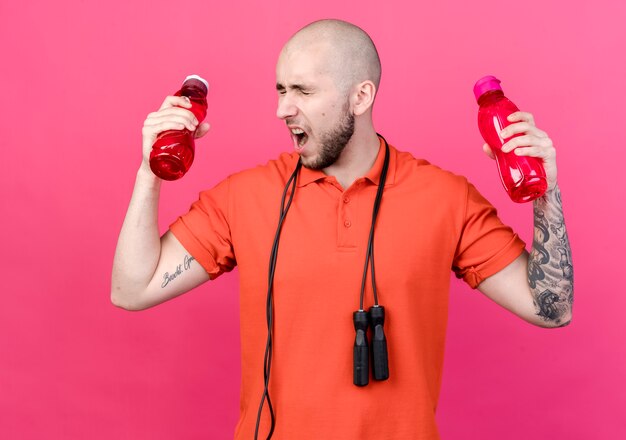 Con los ojos cerrados, enojado, joven deportivo sosteniendo botellas de agua con saltar la cuerda en el hombro aislado en la pared rosa