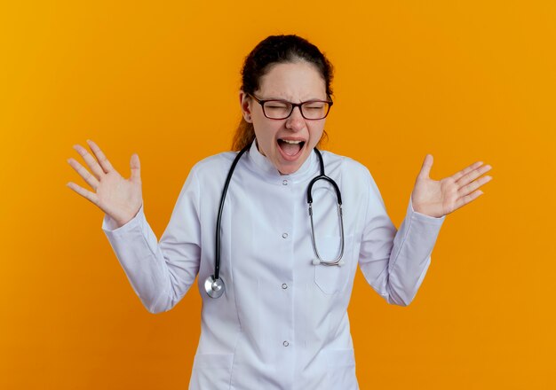 Con los ojos cerrados enojado joven doctora vistiendo bata médica y estetoscopio con gafas extendiendo las manos aisladas en la pared naranja