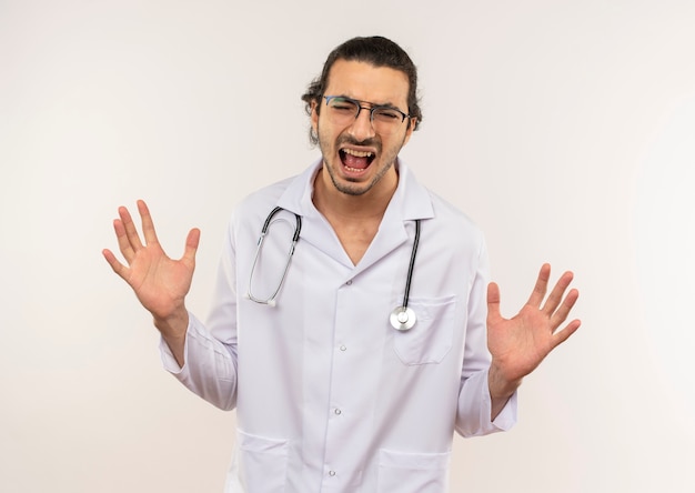 Con los ojos cerrados enojado joven médico con gafas ópticas vistiendo túnica blanca con estetoscopio extiende las manos sobre la pared blanca aislada con espacio de copia