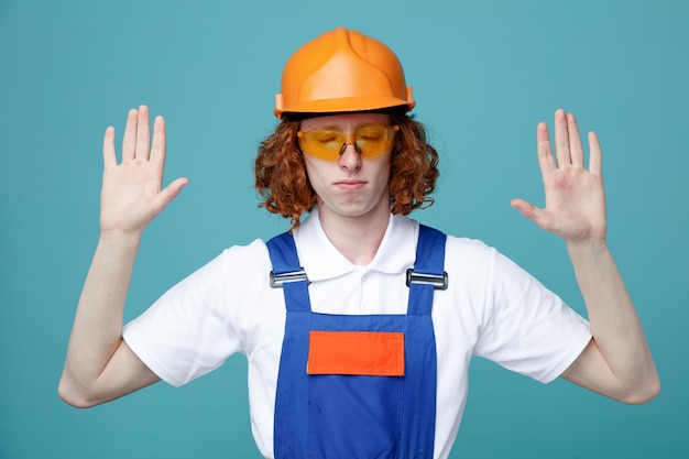 Con los ojos cerrados extendiendo las manos joven constructor hombre en uniforme aislado sobre fondo azul.