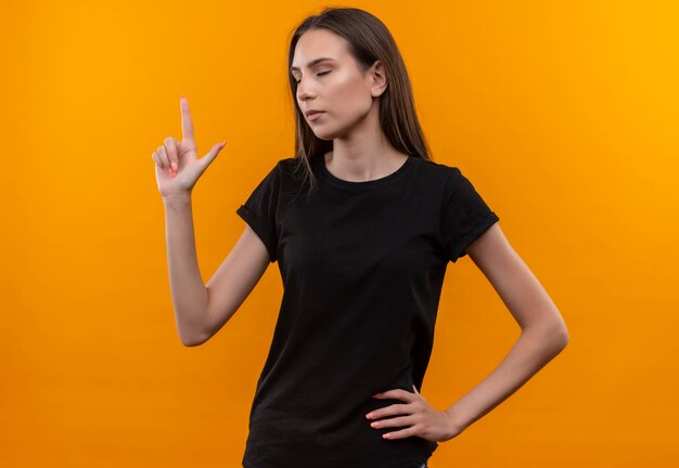 Con los ojos cerrados, joven caucásica con camiseta negra apunta a poner su mano en la cadera en la pared naranja aislada