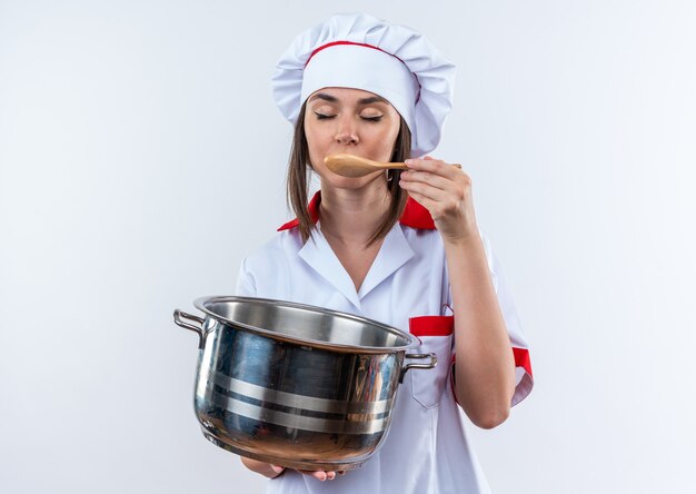 Con los ojos cerrados joven cocinera vistiendo uniforme de chef sosteniendo una cacerola tratando de sopa con cuchara aislado sobre fondo blanco.