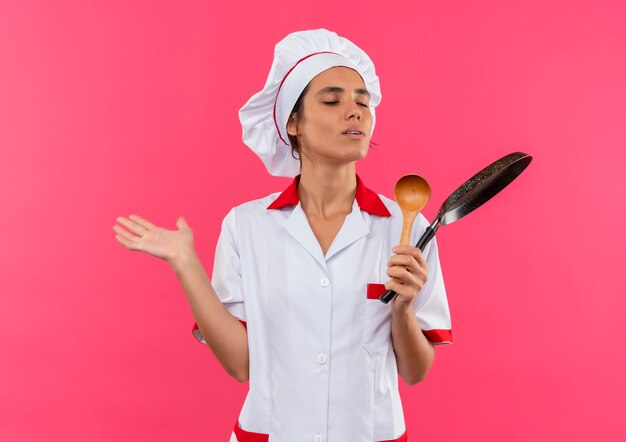 Foto gratuita con los ojos cerrados, joven cocinera vistiendo uniforme de chef sosteniendo una sartén y una cuchara con espacio de copia