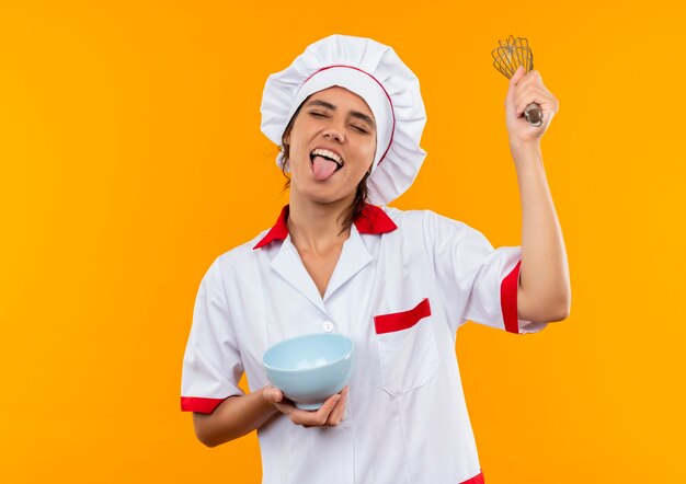 Con los ojos cerrados, joven cocinera vistiendo uniforme de chef sosteniendo el tazón y batir mostrando la lengua con espacio de copia