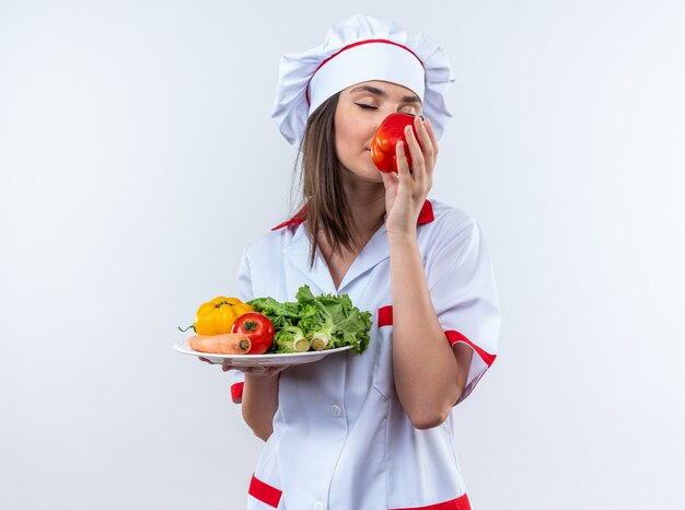 Con los ojos cerrados, joven cocinera vistiendo uniforme de chef sosteniendo verduras en un plato y oliendo pimienta en su mano aislado sobre fondo blanco.