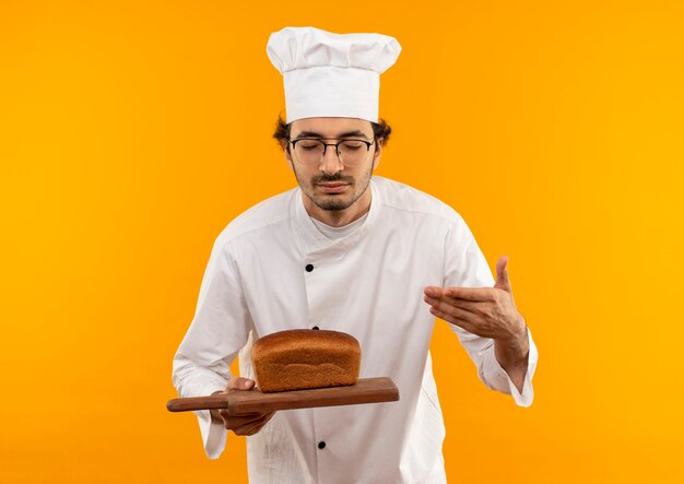 Con los ojos cerrados, joven cocinero con uniforme de chef y gafas sosteniendo y oliendo pan en la tabla de cortar aislada en la pared amarilla