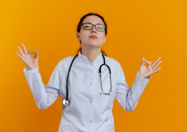 Con los ojos cerrados joven doctora vistiendo bata médica y un estetoscopio con gafas que muestran meditación aislada en la pared naranja