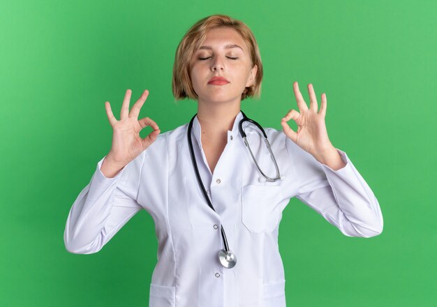 Con los ojos cerrados joven doctora vistiendo bata médica con estetoscopio haciendo meditación aislado sobre fondo verde