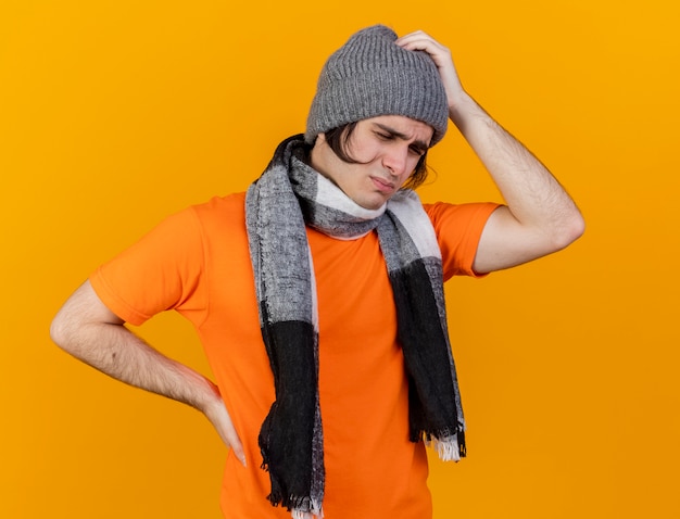 Con los ojos cerrados, joven enfermo disgustado con sombrero de invierno con bufanda poniendo las manos en la cabeza y la cadera doloridas aisladas sobre fondo naranja